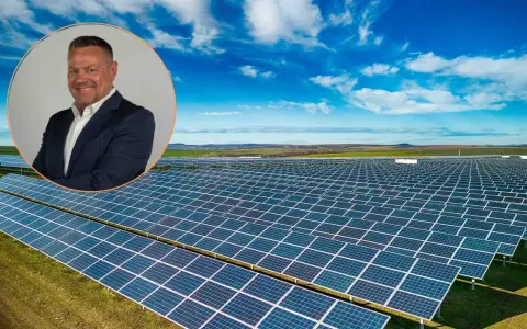 Uśmiechnięty mężczyzna w garniturze na tle farmy fotowoltaicznej. Obraz symbolizuje produkcję energii z fotowoltaiki w poszczególnych miesiącach.