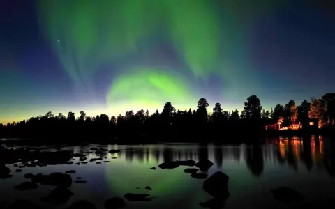 Norrsken dansar över en vinterlandskap vid Kiruna. En skogskant och en sjö med stenar speglar himlens magiska ljus.