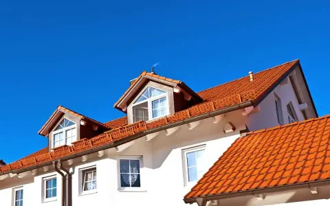 Dach bezokapowy z czerwonej dachówki, z lukarnami i kominem, na tle błękitnego nieba.