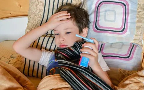 Chory chłopiec leży w łóżku z ręką na czole i trzyma inhalator.