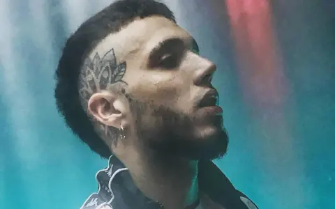 Un joven **hard cantante** con tatuaje en la cabeza y barba, mirando hacia arriba con luces de neón de fondo.