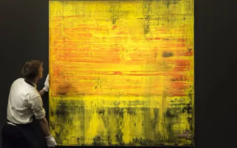 A man in white gloves carefully handles a large abstract painting with vibrant yellow and orange hues, a testament to Gerhard Richter's net worth.