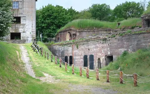 Fort Zachodni w Świnoujściu, zarośnięty trawą i otoczony zielenią, z widocznym starym budynkiem i historyczną artylerią.