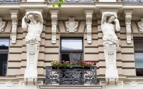 Dwie rzeźby atlantesów podtrzymujące balkon, przykład secesji w sztuce, zdobią fasadę kamienicy.