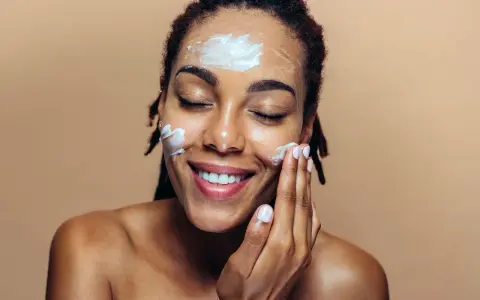 Une femme applique de la crème sur son visage. Elle sourit, les yeux fermés. Elle se demande que veut dire indice 50 pour sa protection solaire.
