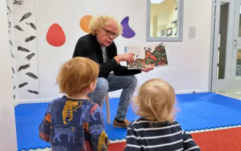 En man med lockigt hår läser en bok för två små barn. Bilden visar en humlan djojj på en morot i boken.