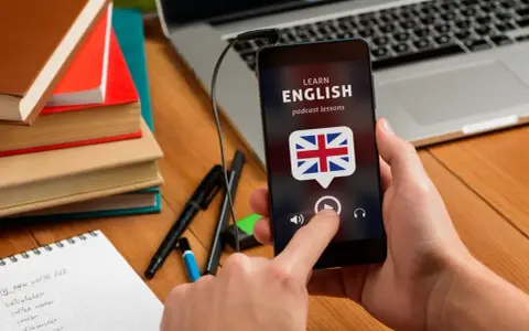 Ręka trzyma smartfon z aplikacją do nauki angielskiego. Na biurku leżą książki i notatnik z nowymi słówkami.
