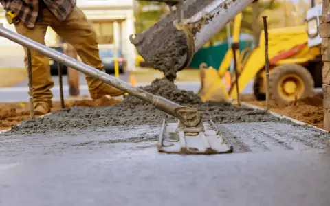 Pracownik wylewa beton, który będzie schnął. W tle koparka i budowa.