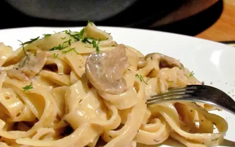 Tagiatelle z grzybami w kremowym sosie, posypane świeżym koperkiem. Widok z bliska na talerz pełen pysznego makaronu.