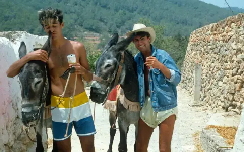Tony Pike Ibiza: due ragazzi con asini, uno con pantaloncini colorati, l'altro con giacca di jeans e cappello di paglia, sorseggiano bevande.