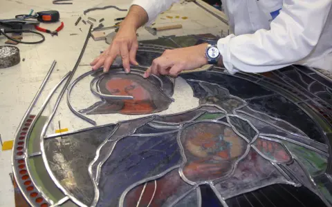 Un artisan assemble un vitrail, joignant des pièces de verre coloré avec du plomb. Le visage d'une Vierge et d'un enfant se dessine.