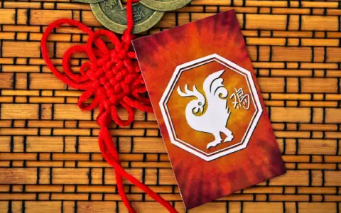 Czerwony chiński węzeł i karta z białym kogutem, symbolem 1993 chińskiego znaku zodiaku, na tle bambusowym.