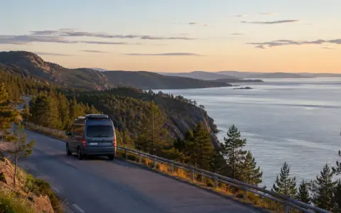 En husbil kör längs en slingrande väg vid den vackra Höga kusten. Solnedgången färgar himlen i mjuka toner över det glittrande havet.