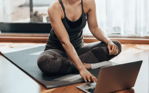 Kobieta w stroju sportowym siedzi na macie, korzystając z laptopa. Czy można ćwiczyć codziennie? Tak, jeśli masz dostęp do inspiracji online!