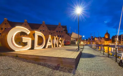 Podświetlany napis "GDAŃSK" na tle nocnej panoramy miasta, zapraszający do odkrywania atrakcji Trójmiasta.
