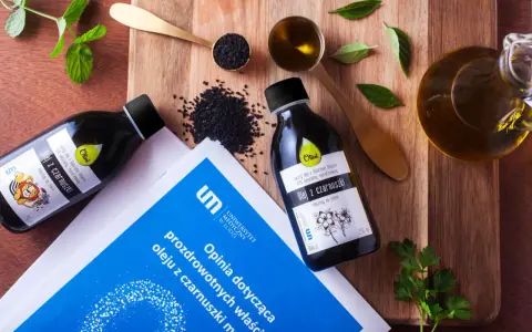 Olej z czarnuszki, bogaty w kwasy omega-3, prezentowany na desce z nasionami i ziołami. Obok butelki z olejem leży dokument z opinią o jego właściwościach.