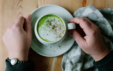 Händerna håller en kopp med en krämig matcha latte, toppad med skum och lite kryddor. En mysig stund med en grön dryck.