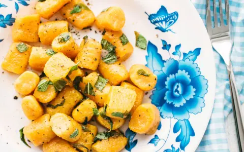 Gnocchi di zucca dorati, conditi con erbe aromatiche e pepe nero, su un piatto decorato con fiori blu.