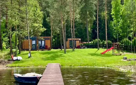 Drewniany pomost nad jeziorem Bardze, z łódką przycumowaną do brzegu. W tle domki letniskowe i plac zabaw.