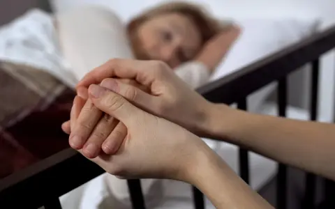 Dwie dłonie splatają się, jedna z nich spoczywa na ramieniu śpiącej osoby. To obraz towarzyszenia w trudnych chwilach, gdy rozważana jest eutanazja co to.