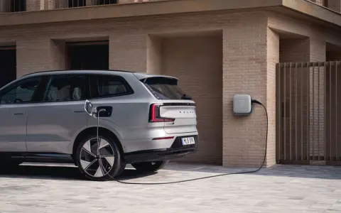 Srebrny, duży SUV Volvo EX90 podłączony do ładowarki ściennej. Nowoczesny design i ekologiczna energia.