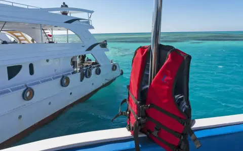Gilet de sauvetage rouge prêt pour l'aventure en mer, à côté d'un yacht blanc. Les **œuvres mortes du bateau** se reflètent dans l'eau turquoise.