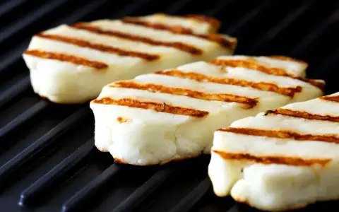 Trzy kawałki halloumi z grilla, z apetycznymi paskami, leżą na czarnej grillowej płycie.