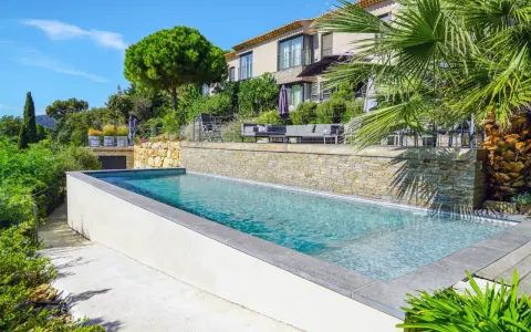 Belle piscine semi enterrée avec un habillage en pierre naturelle, entourée de verdure luxuriante et d'une villa moderne.