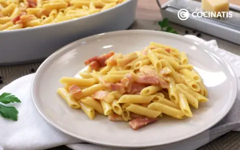 Plato de macarrones a la carbonara cremosos con trozos de panceta. En el fondo, un bol con más pasta y un rallador de queso.