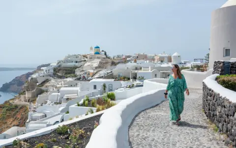 Kobieta w zwiewnej sukience spaceruje po Santorini. Idealne miejsce, gdzie do Grecji na wakacje, z widokiem na białe domy i błękitne kopuły.
