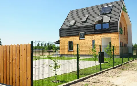 Drewniany płot i siatka ogrodzeniowa przed nowoczesnym domem z panelami słonecznymi.