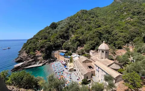 Baia con spiaggia affollata e un antico monastero, un luogo ideale per i cammini in Liguria, con mare azzurro e vegetazione rigogliosa.