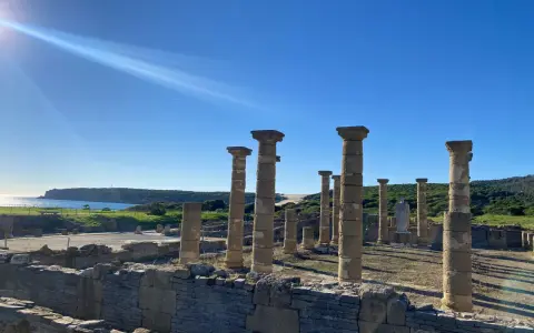 Sunlight streams over ancient roman ruins in Spain, illuminating weathered stone columns and walls against a bright blue sky and distant coastline.