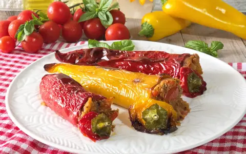 Peperoni cornetti ripieni, rossi e gialli, cotti al forno, su un piatto bianco con pomodorini e basilico.