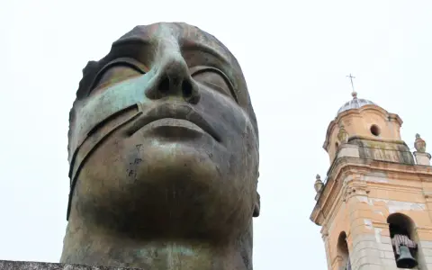 Brązowa, monumentalna rzeźba głowy z widocznym fragmentem dzwonnicy kościelnej w tle.