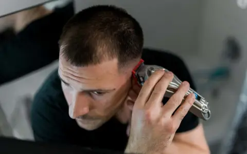 Mann mit Haarschneidemaschine am Hinterkopf. Er schneidet sich die Haare selber, wie eine Frau, die ihre Haare zu Hause schneidet.