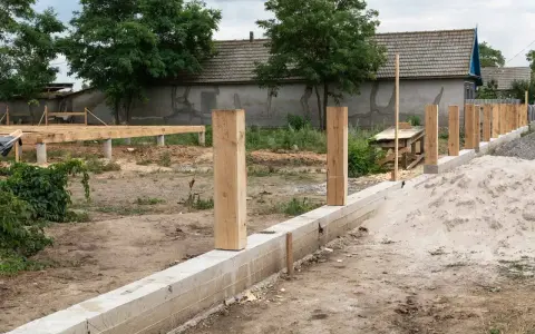Solidny fundament pod ogrodzenie z drewnianymi słupkami, w tle budowa domu.