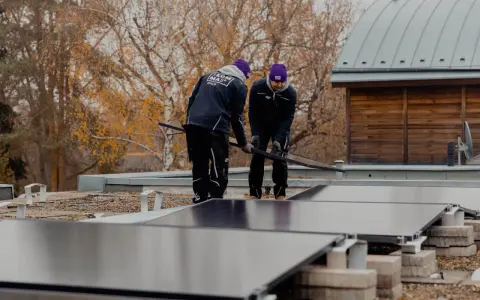 Två personer installerar solceller på ett papptak. Höstfärgade träd syns i bakgrunden.