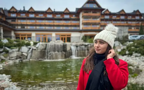 Kobieta w czerwonym płaszczu i białej czapce przed hotelem Bania. W tle wodospad i staw. Wiele pozytywnych hotel Bania opinie potwierdza ten widok.