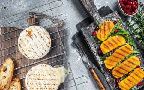 Dwa okrągłe serki na grilla, jeden lekko roztopiony, obok pieczywo. Na desce grillowane kawałki dyni z granatem i rukolą.