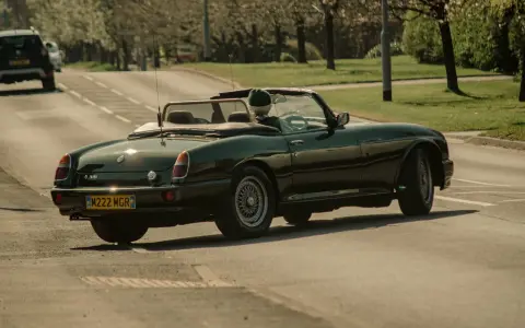Auto d'epoca verde scuro in movimento, con conducente con cappello. La targa suggerisce che siamo in Inghilterra, dove si guida a destra.