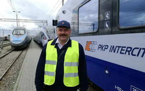 Uśmiechnięty rewident taboru kolejowego w kamizelce odblaskowej stoi obok pociągu PKP Intercity.