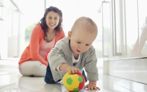 Maluch ćwiczy motorykę dużą, raczkując po podłodze i bawiąc się kolorową piłeczką. Uśmiechnięta mama obserwuje jego postępy.