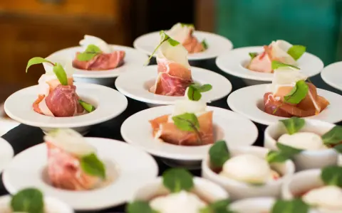 Eleganckie przekąski na białych talerzykach, zawinięte w szynkę parmeńską z listkami ziół. To właśnie catering, który zachwyca smakiem i wyglądem.