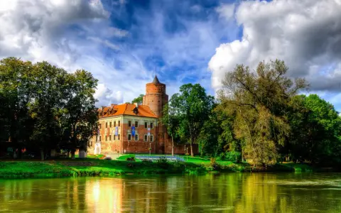 Malowniczy zamek w Łódzkiem nad rzeką, otoczony zielenią i chmurami.