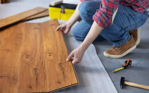 Persona instala tarima flotante, uniendo tablas de madera con cuidado. Herramientas como martillo y cinta métrica a mano.