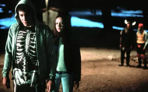 Donnie Darko w bluzie z kapturem i koszulce z nadrukiem szkieletu, obok dziewczyny. W tle dwie postacie w kostiumach.