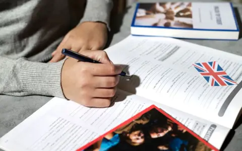 Osoba pisze ćwiczenia z angielskiego, obok leży książka i flaga Wielkiej Brytanii. Klauzula CV po angielsku może być częścią takich materiałów.
