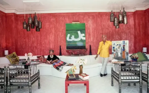 Interni di case anni '70: una donna legge sul divano bianco, un uomo mostra un quadro. Pareti rosse, lampade a grappolo.