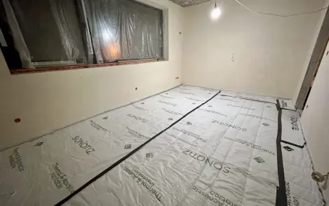 W trakcie prac remontowych w bloku, podłoga pokryta jest materiałem do wyciszenia. Widać okno zabezpieczone folią i sufit z widocznymi instalacjami.